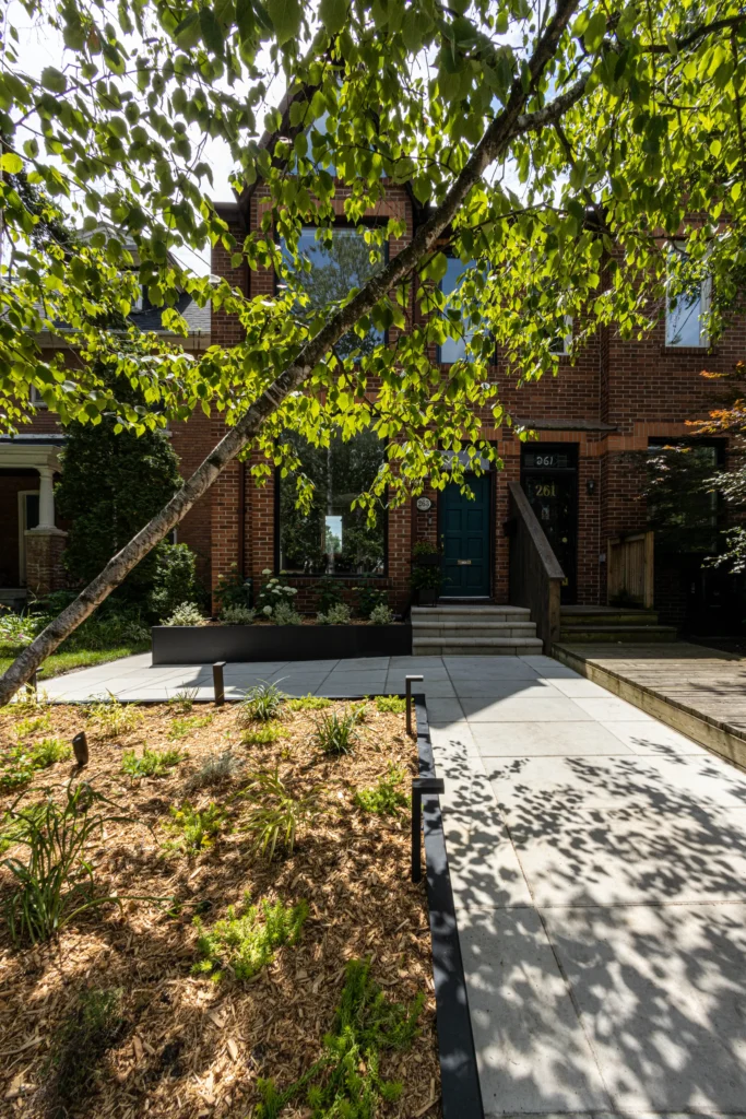 Front yard landscaping in Toronto with raised planter bed, mature tree, modern stone walkway, and soft landscape lighting in front of brick home