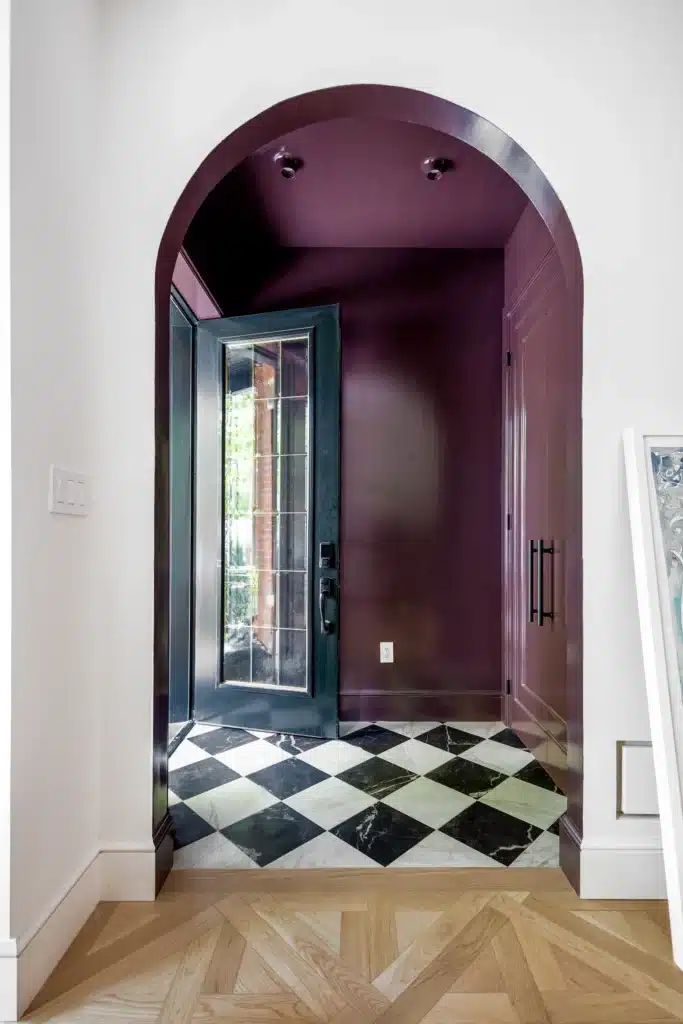 Renovated home entryway with a deep burgundy arched alcove, black glass front door, black-and-white checkerboard floor, and light wood flooring.