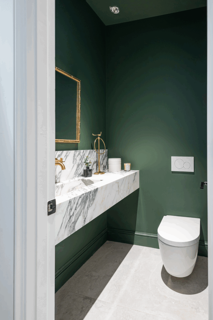 Renovated powder room with dark green walls, a marble vanity, brass fixtures, and a wall-mounted toilet.