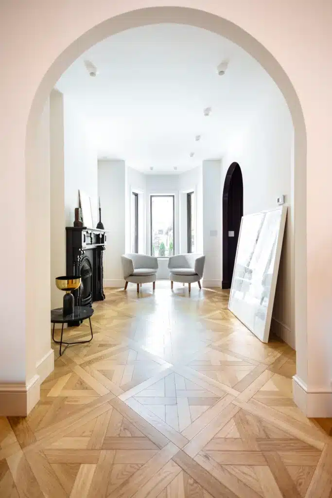 Renovated home interior with a wide arched doorway, patterned wood floors, a black fireplace, and two chairs beside a sunlit window.