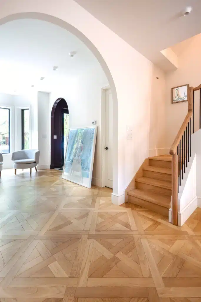 Renovated home interior with a wide arched doorway, patterned wood floors,, and chairs beside a sunlit window.