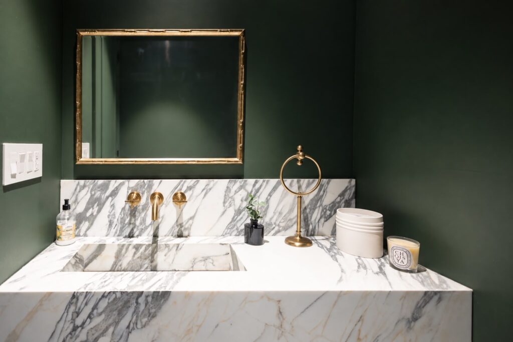 Renovated powder room with dark green walls, a marble vanity, brass fixtures, and a wall-mounted toilet.