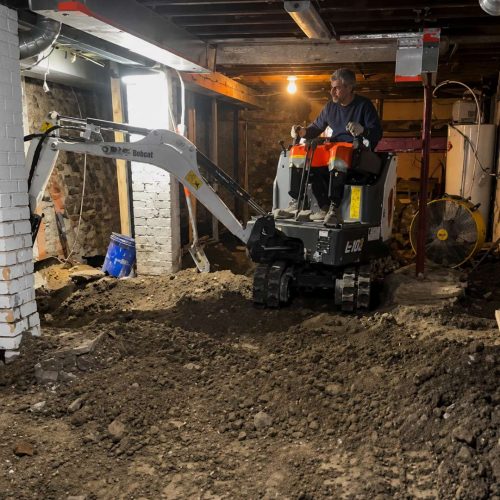 Mini excavator operating inside a residential basement during underpinning work with exposed soil, low ceilings, and active excavation in Toronto home renovation project.