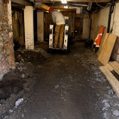 Skid steer excavator operating inside a Toronto basement during underpinning and basement lowering construction with exposed foundation walls and excavated soil.